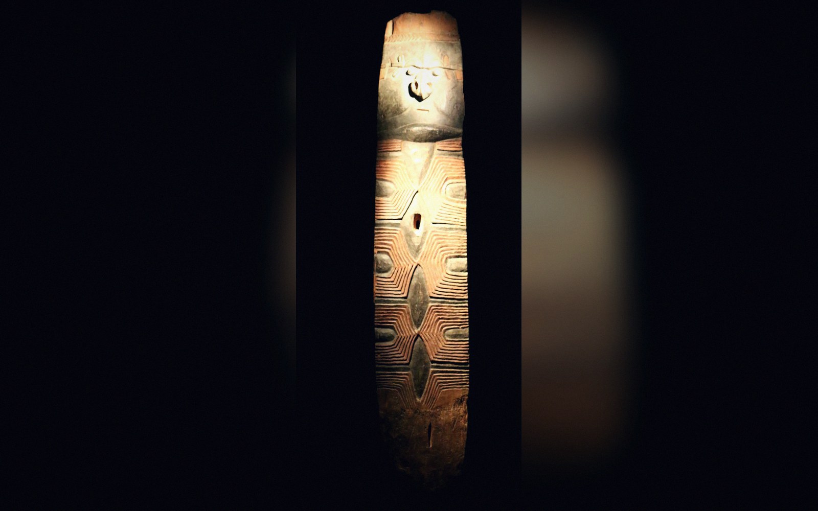 Carved wooden post with a stylized face and intricate geometric line patterns, lit against darkness